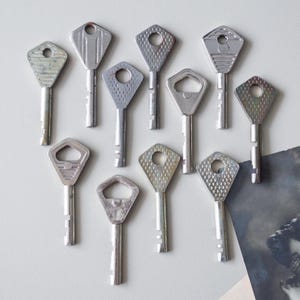 May include: A collection of vintage silver keys with diamond-shaped heads. The keys have various textured patterns and circular or keyhole-shaped openings. The keys are arranged on a light gray surface, suggesting a focus on antique or collectible items.