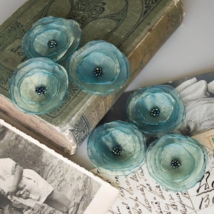Könnte beinhalten: Sechs teal-farbene Stoffblumen-Verzierungen mit Perlen in der Mitte. Die Blumen sind mit einem transparenten, strukturierten Material geschichtet. Sie sind auf einem Vintage-Buch und alten Fotografien angeordnet.