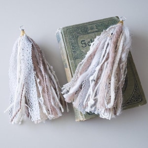 May include: Two white and brown yarn tassels with white lace accents. The tassels are attached to silver rings and are hanging from a vintage book.