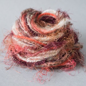 Puede incluir: Un ovillo de lana en tonos rojos, blancos y marrones. La lana es muy suave y tiene mucha textura.