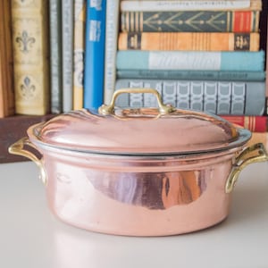Peut inclure: Une casserole ovale en cuivre avec des poignées en laiton et un couvercle. Le plat est posé sur une surface blanche.