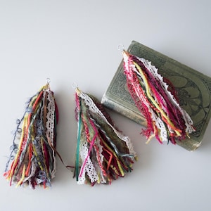 Puede incluir: Tres borlas de hilo de colores con detalles de encaje blanco y tapas doradas. Las borlas cuelgan de un libro.