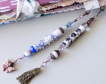 May include: Two handmade beaded bookmarks. One features a butterfly charm, the other a tassel. Both have decorative beads in blue, white, and clear, with metal accents. The bookmarks are designed for journals or books.