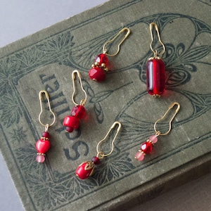 May include: Six handcrafted stitch markers with gold-colored safety pin clasps. Each marker features a unique design with red and pink beads, including faceted crystals and round beads. The markers are displayed on a vintage book.
