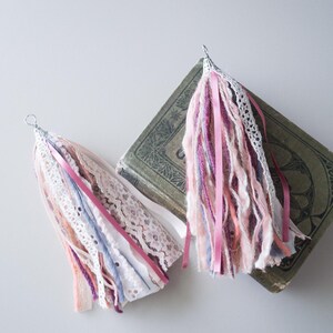 May include: Two pink, white and grey yarn tassels with a silver metal ring at the top. The tassels are made of yarn, lace and ribbon.