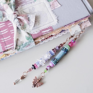 Puede incluir: Dos colgantes de marcapáginas con cuentas y cierres de color cobre. Uno presenta un dije de mariposa, el otro un cristal transparente. Los marcapáginas están junto a un diario con un lazo de tela floral y una cubierta de patchwork.