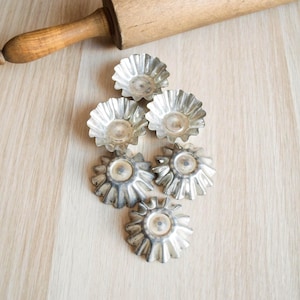 May include: A wooden rolling pin lies diagonally across a light-coloured wooden surface. Below it are six small, silver, fluted tart or cupcake tins arranged in a cluster. The tins have a vintage, slightly tarnished appearance, suggesting age and use.