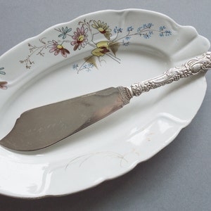 May include: A silver butter knife with an ornate handle rests on a white oval serving dish with a floral pattern. The dish has a scalloped edge and is decorated with blue, pink, and yellow flowers.