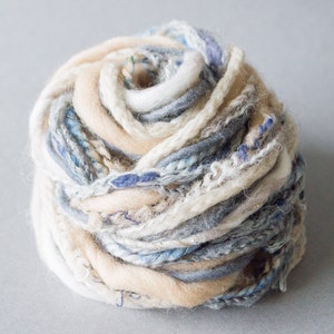 Może przedstawiać: Kulka włóczki w mieszance kolorów niebieskiego, białego i beżowego. Włóczka jest wykonana z różnych faktur i ma puszysty wygląd.