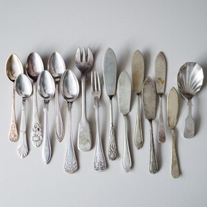 May include: A collection of vintage silverware, including spoons, forks, and butter knives, arranged in a row on a white surface. The silverware is made of silver and has intricate designs.