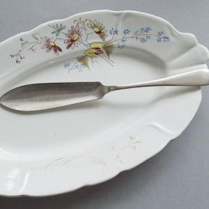 May include: A white oval serving dish with a floral design in shades of blue, yellow, pink, and brown. A silver butter knife is resting on the dish.