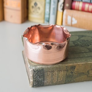 Könnte beinhalten: Eine kupferfarbene Schale mit gewelltem Rand und polierter Oberfläche. Die zylindrische Schale hat ein dekoratives horizontales Band. Sie liegt auf einem Vintage-Buch mit verblasstem grünem Einband und goldener Schrift. Bücher sind im Hintergrund.