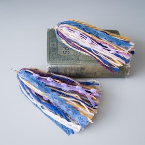 May include: Two colourful tassel keychains with a mix of blue, yellow, purple, and white yarn, lace, and ribbon. The tassels are hanging from a silver ring.