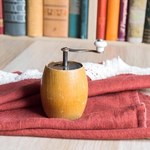 以下が含まれることがあります： 金属製のクランクと白いセラミック製のハンドルが付いた、ヴィンテージの木製ペッパーグラインダー。 グラインダーは円筒形で、白いレースのトリムが付いた赤い布の上に置かれています。 背景には本棚があります。