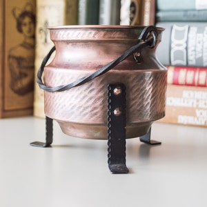 May include: A copper cauldron with a black iron handle and three black iron legs. The cauldron has a hammered texture and a rustic finish.