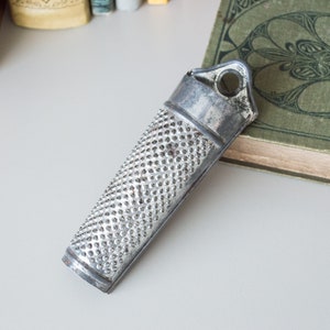 Op de afbeelding: Een zilveren metalen rasp met een handvat en een gat om op te hangen. De rasp heeft een getextureerd oppervlak met kleine gaatjes.