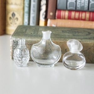 May include: Three clear glass perfume bottles of varying shapes and sizes. The bottles are empty and sit on a white surface in front of a stack of antique books. One bottle has a textured, patterned design.