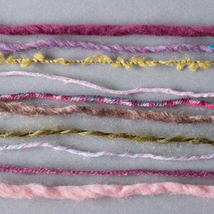 May include: A collection of ten different colored yarn strands, including pink, purple, brown, and green, are laid out horizontally on a gray surface.