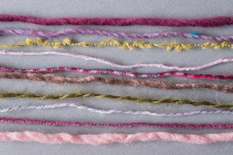May include: A collection of ten different colored yarn strands, including pink, purple, brown, and green, are laid out horizontally on a gray surface.