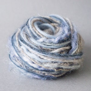 Puede incluir: Un ovillo de lana en tonos azules, blancos y beige. La lana es suave y esponjosa, y es perfecta para tejer o hacer ganchillo.