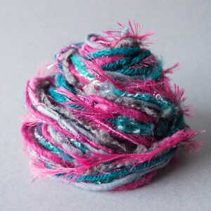 Puede incluir: Una bola de hilo con hebras rosas, azul verdoso y grises. El hilo es esponjoso y tiene un aspecto texturizado.