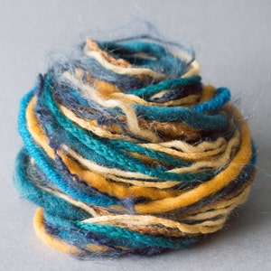 Puede incluir: Un ovillo de hilo en tonos de azul verdoso, azul, marrón y crema. El hilo está retorcido y apilado en forma de bola.