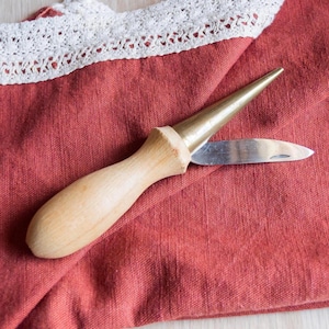 May include: A leatherworking tool with a wooden handle and a brass tip, along with a small, curved blade. The tool rests on a red fabric, with a white lace trim visible in the background. This tool is likely used for crafting or leatherwork.
