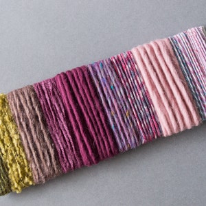 May include: A collection of colorful yarn strands in shades of green, brown, pink, purple, and white. The yarn is arranged in a row on a white surface.