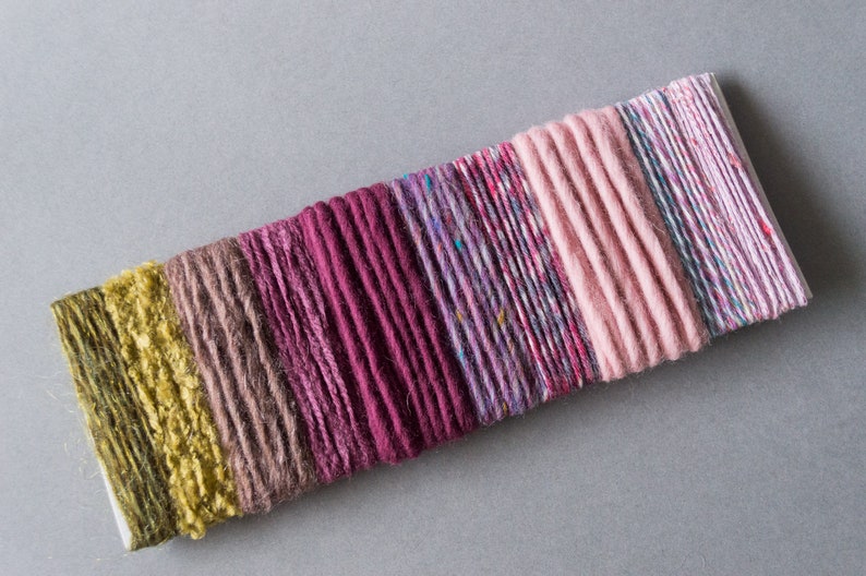 May include: A collection of colorful yarn strands in shades of green, brown, pink, purple, and white. The yarn is arranged in a row on a white surface.