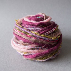 May include: A ball of yarn with multiple strands of pink, purple, brown, and gold yarn. The yarn is loosely wound and has a rustic, handmade look.