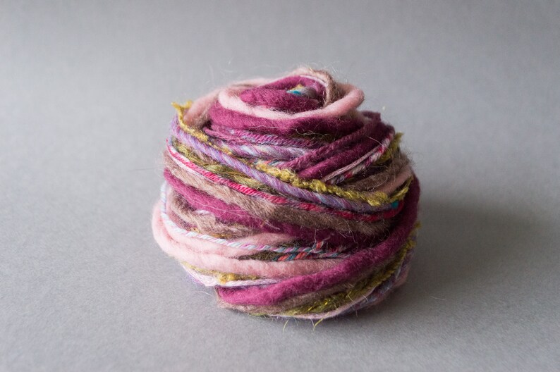 May include: A ball of yarn with multiple strands of pink, purple, brown, and gold yarn. The yarn is loosely wound and has a rustic, handmade look.
