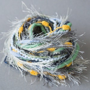 Puede incluir: Primer plano de un ovillo de hilo en tonos blancos, grises, verdes y amarillos. El hilo es esponjoso y texturizado, con pequeños pompones amarillos por todas partes.