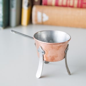 May include: A small copper pot with a silver handle and three silver legs. The pot is sitting on a white surface.
