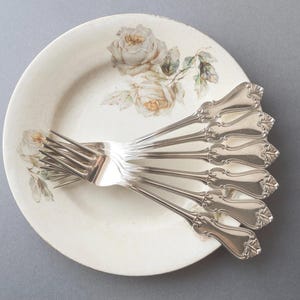 May include: A set of six silver-toned metal forks with ornate, flared handles arranged in a fan shape on top of a round, off-white ceramic plate decorated with a delicate, muted-colour floral pattern featuring pale roses and green leaves.