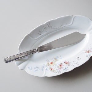 May include: A white oval serving dish with a floral pattern in shades of pink, blue, and yellow. A silver butter knife is placed on the dish.