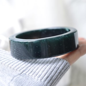 May include: A smooth, polished bangle bracelet in a deep, dark forest green color. The circular accessory features a solid, thick band with subtle, lighter green speckled inclusions throughout the polished surface, creating a natural appearance.