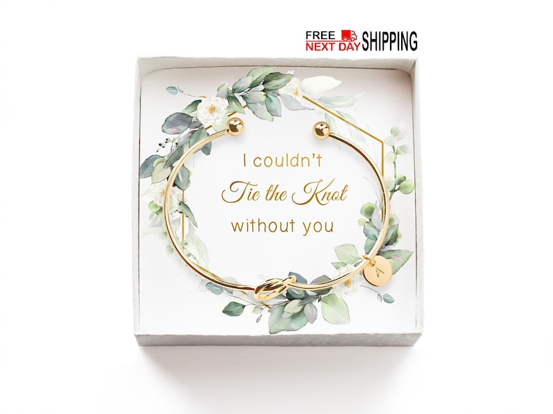 A knot bracelet rests on a white surface. The bracelet has a polished band forming a simple knot at the center, with a small, round charm engraved with an initial hanging from the knot. The design is elegant and minimalist.