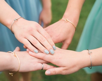May include: Four hands stacked on top of each other, each wearing a simple gold or silver knot bracelet. The hands have light blue nail polish.