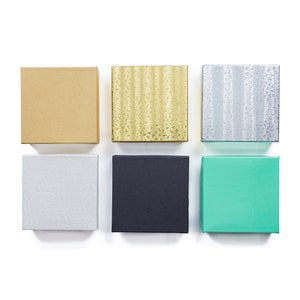 May include: Six small gift boxes in various colors and textures. The boxes are arranged in two rows of three. The top row features a brown box, a gold box with a textured pattern, and a silver box with a textured pattern. The bottom row features a white box with a textured pattern, a black box, and a teal box.