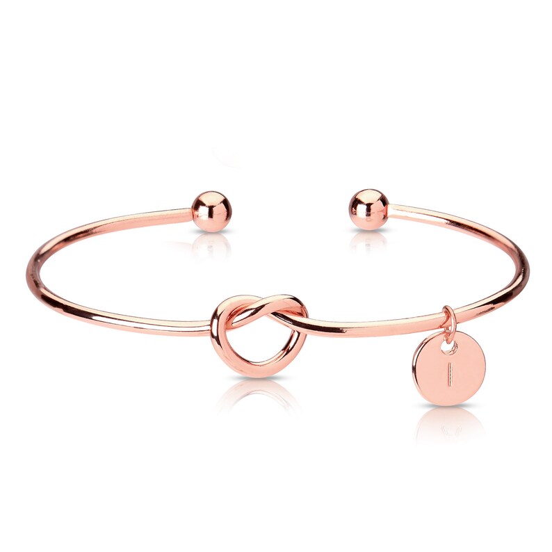May include: Rose gold knot bangle bracelet with a small round charm with the letter I.