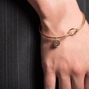 May include: A gold bangle bracelet with a knot design and a small round charm with the letter "I" on it.
