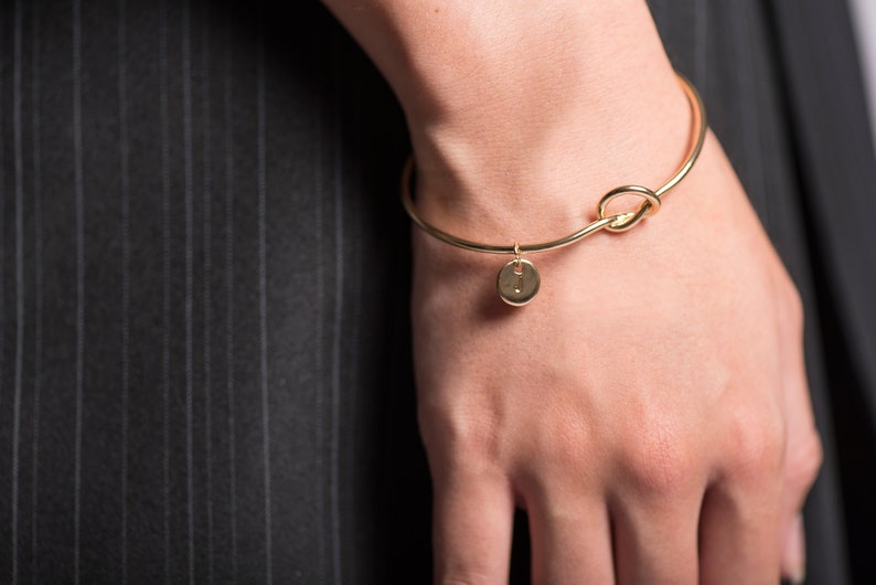 May include: A gold bangle bracelet with a knot design and a small round charm with the letter "I" on it.