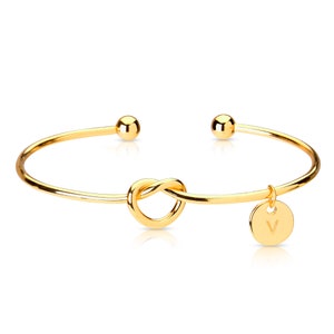 May include: Gold bangle bracelet with a knot design and a small charm with the letter 'V' hanging from it.