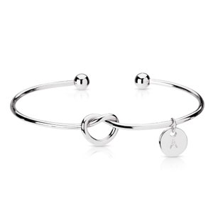 May include: Silver knot bangle bracelet with a small charm that has the letter 'A' on it.