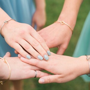 May include: Four hands stacked on top of each other, each wearing a simple gold or silver knot bracelet. The hands have light blue nail polish.