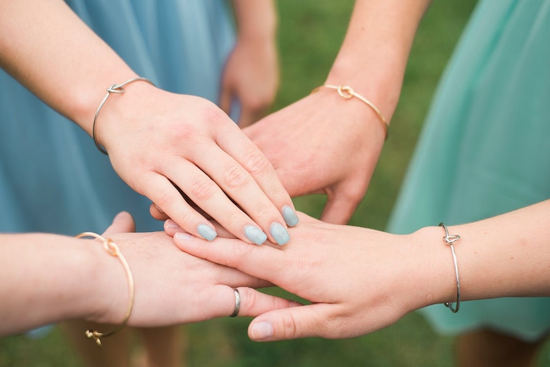 May include: Four hands stacked on top of each other, each wearing a simple gold or silver knot bracelet. The hands have light blue nail polish.