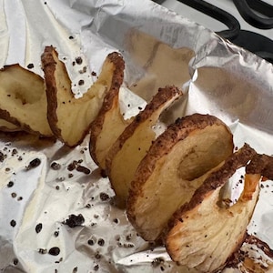 Puede incluir: Una espiral de rodajas de patata, fritas a un dorado marrón, en una bandeja de horno forrada con papel de aluminio plateado. La patata está sazonada con pimienta negra.