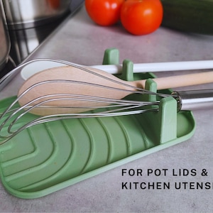 May include: Green silicone kitchen utensil holder with a textured surface. It has slots for holding wooden spoons, whisks, and other kitchen tools. The text "FOR POT LIDS & KITCHEN UTENSILS" is printed on the holder.