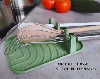May include: Green silicone kitchen utensil holder with a textured surface. It has slots for holding wooden spoons, whisks, and other kitchen tools. The text "FOR POT LIDS & KITCHEN UTENSILS" is printed on the holder.
