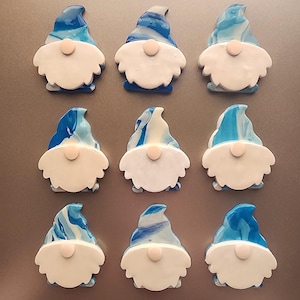 May include: Nine gnome-shaped magnets. Each gnome has a white beard, a pink nose, and a blue and white swirled hat. The magnets are arranged in three rows of three.